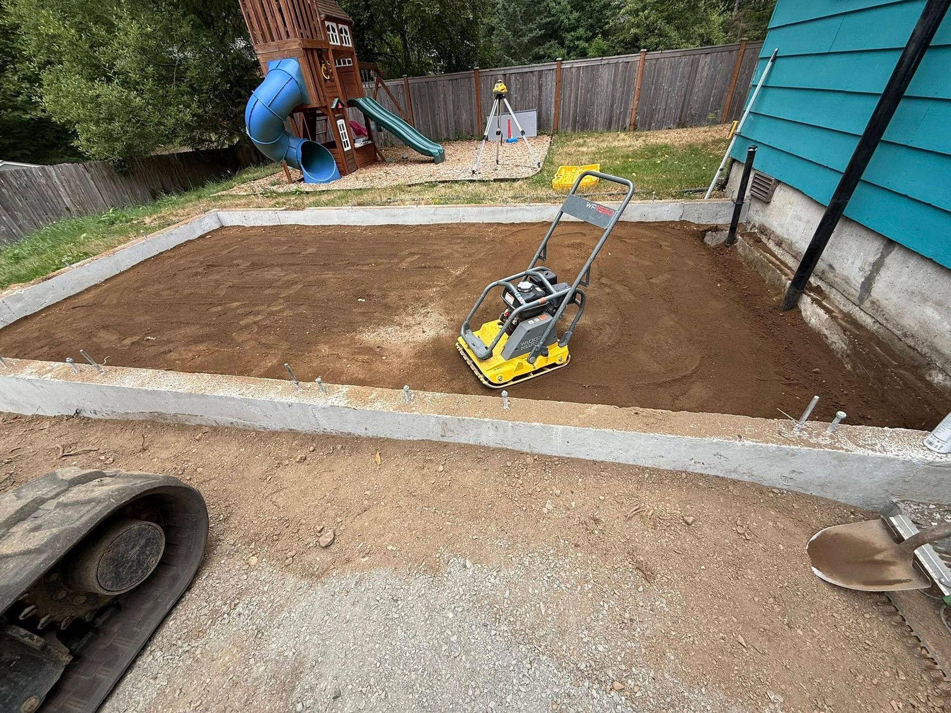 A yellow plate compactor sits inside a rectangular concrete foundation filled with soil in a backyard.