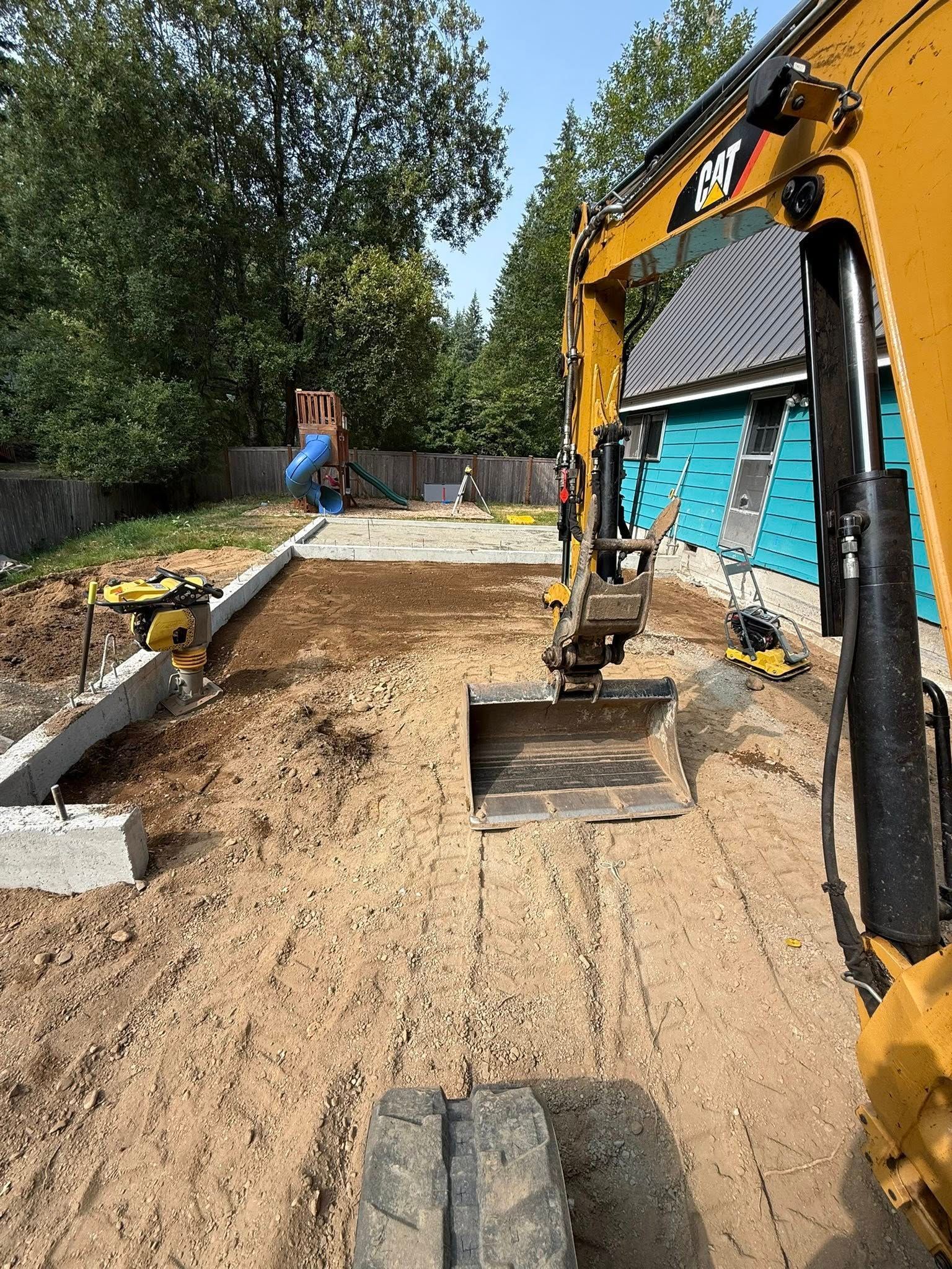 An excavator with a bucket sits in a backyard on a dirt lot beside a partially constructed concrete foundation.
