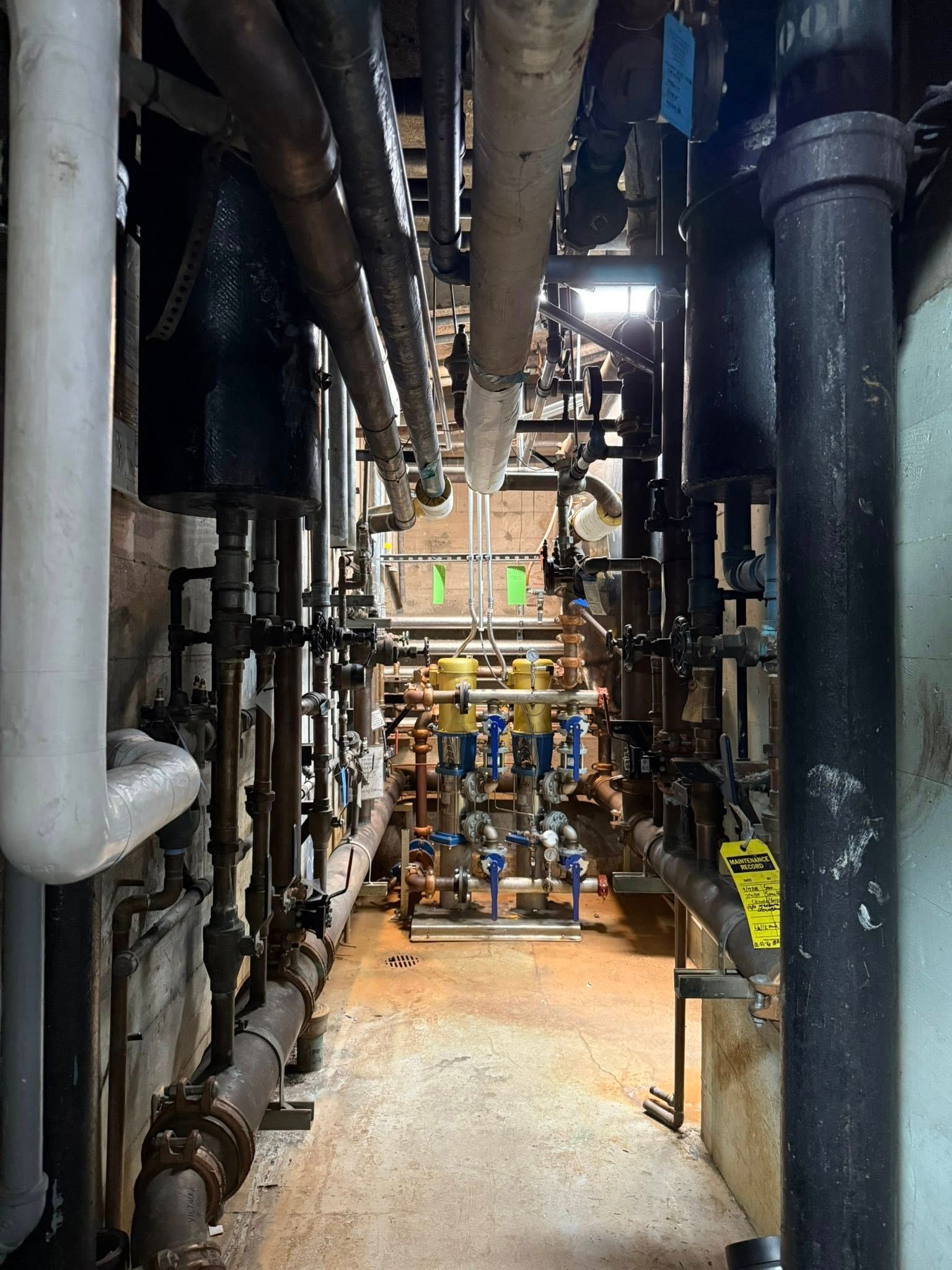 A narrow industrial utility corridor lined with various metal pipes and valves, illuminated by a light at the far end.