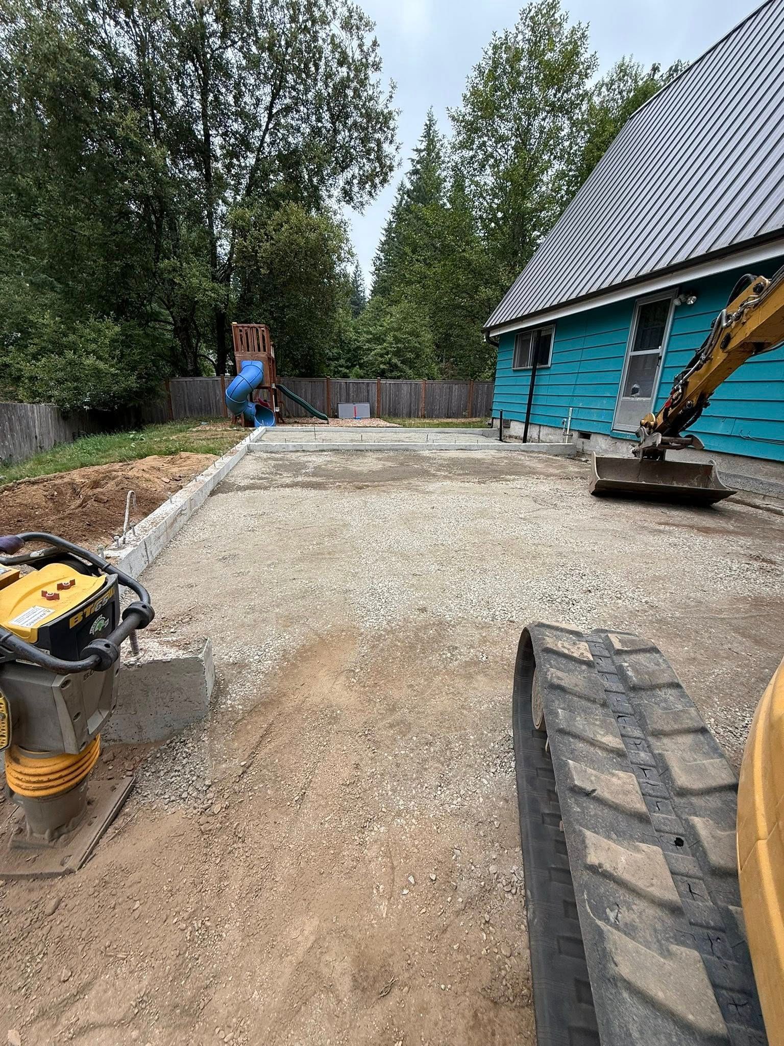 A construction site with a teal house, a gravel patio base, and heavy machinery, including an excavator.