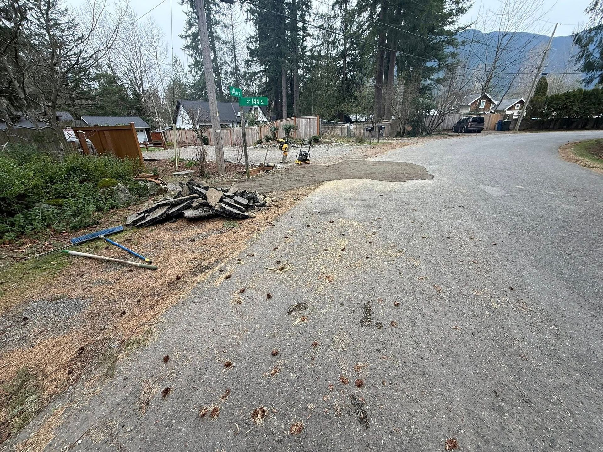 A gravel road curves past a patch of dirt and debris in a wooded, residential area with a green street sign in the distance.