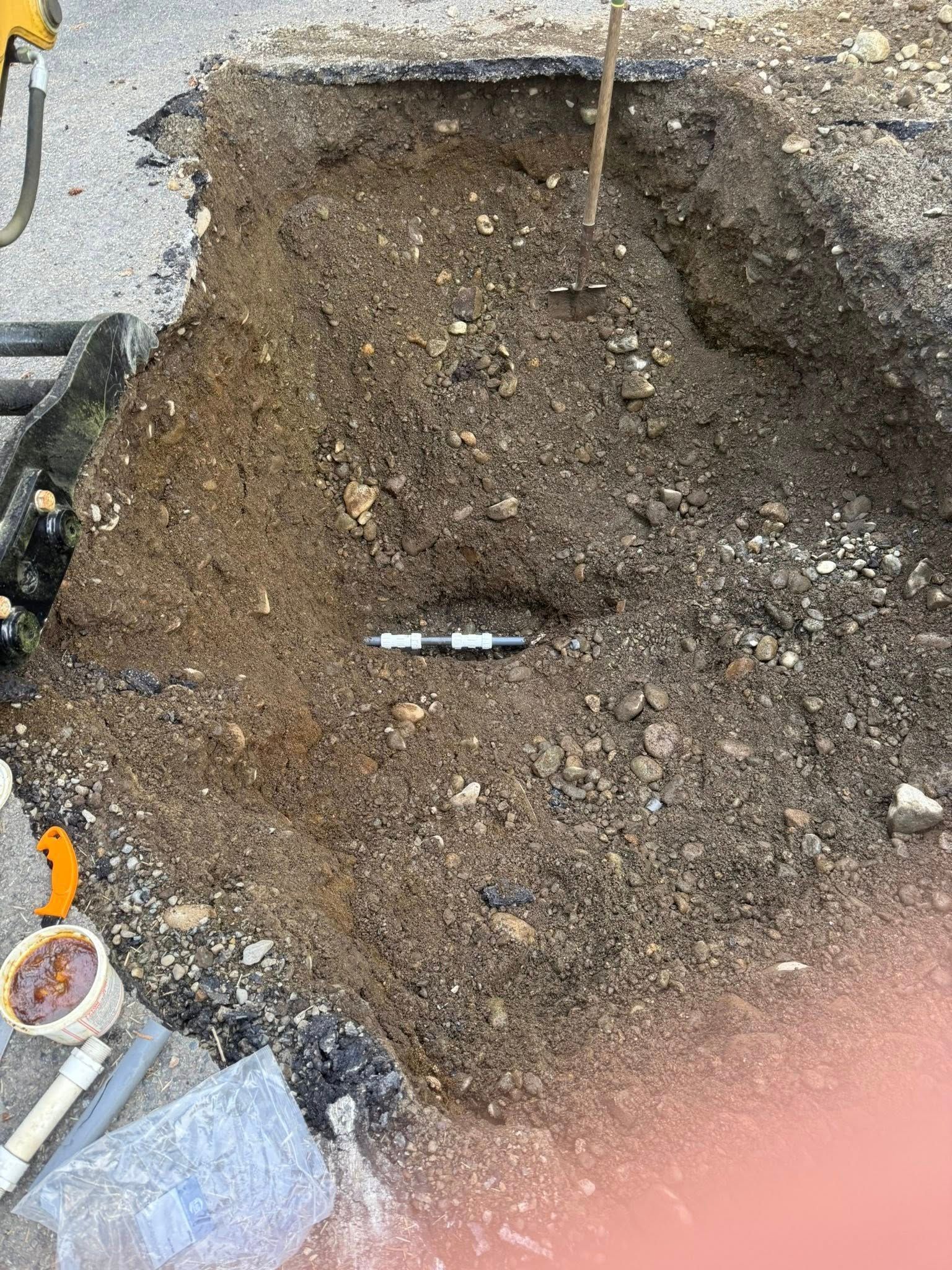 A rectangular excavation pit in asphalt, showing exposed dirt, a small section of black pipe, and construction equipment.