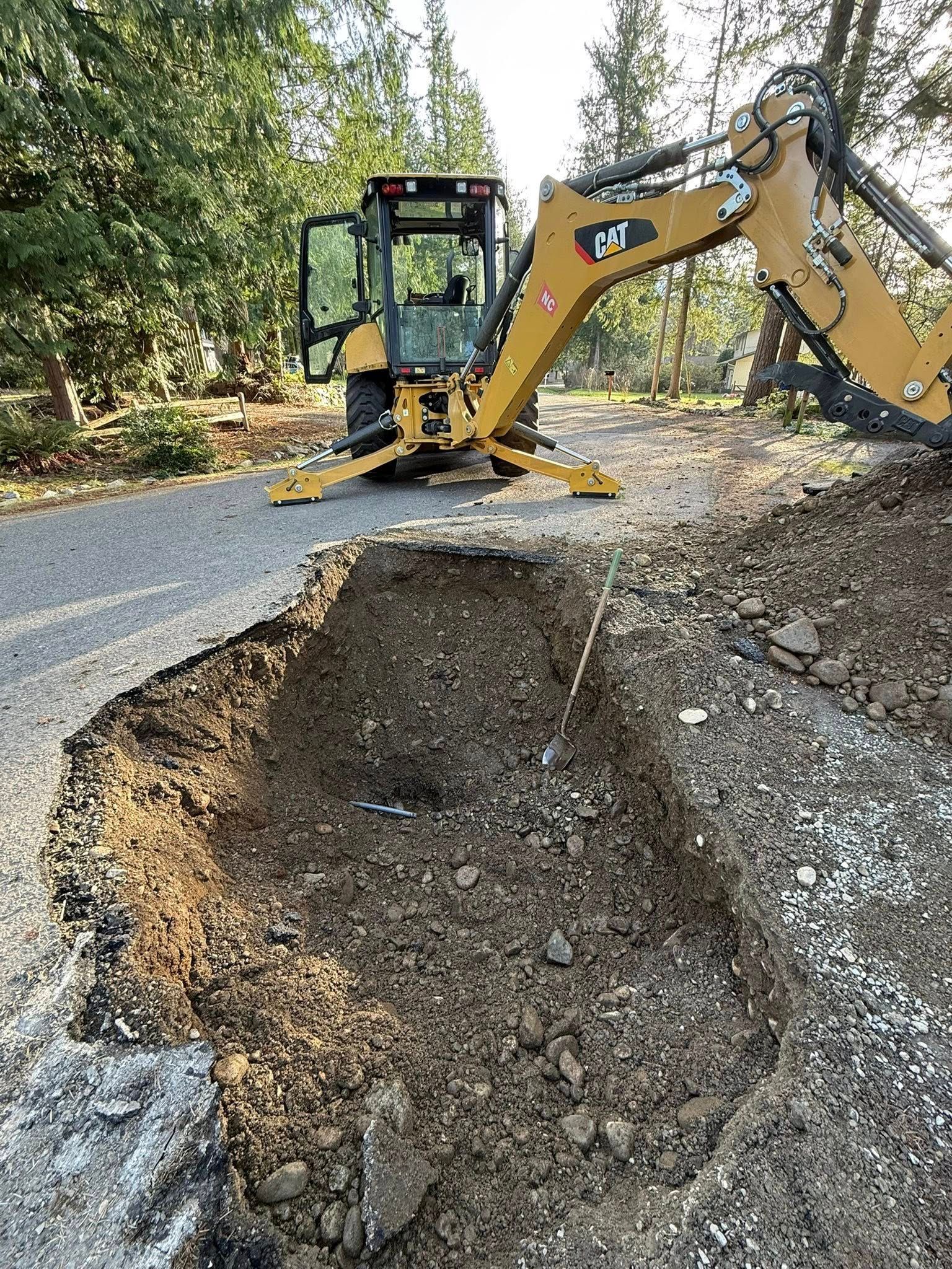 A yellow Caterpillar backhoe parked on a paved road next to a large trench excavated in the dirt.