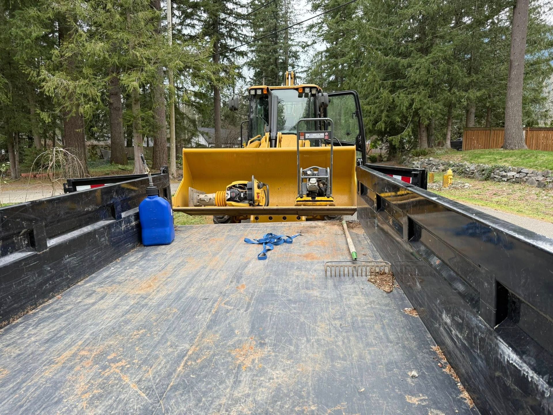 A yellow tractor bucket sits on a flatbed trailer containing a blue fuel jug and a metal rake.