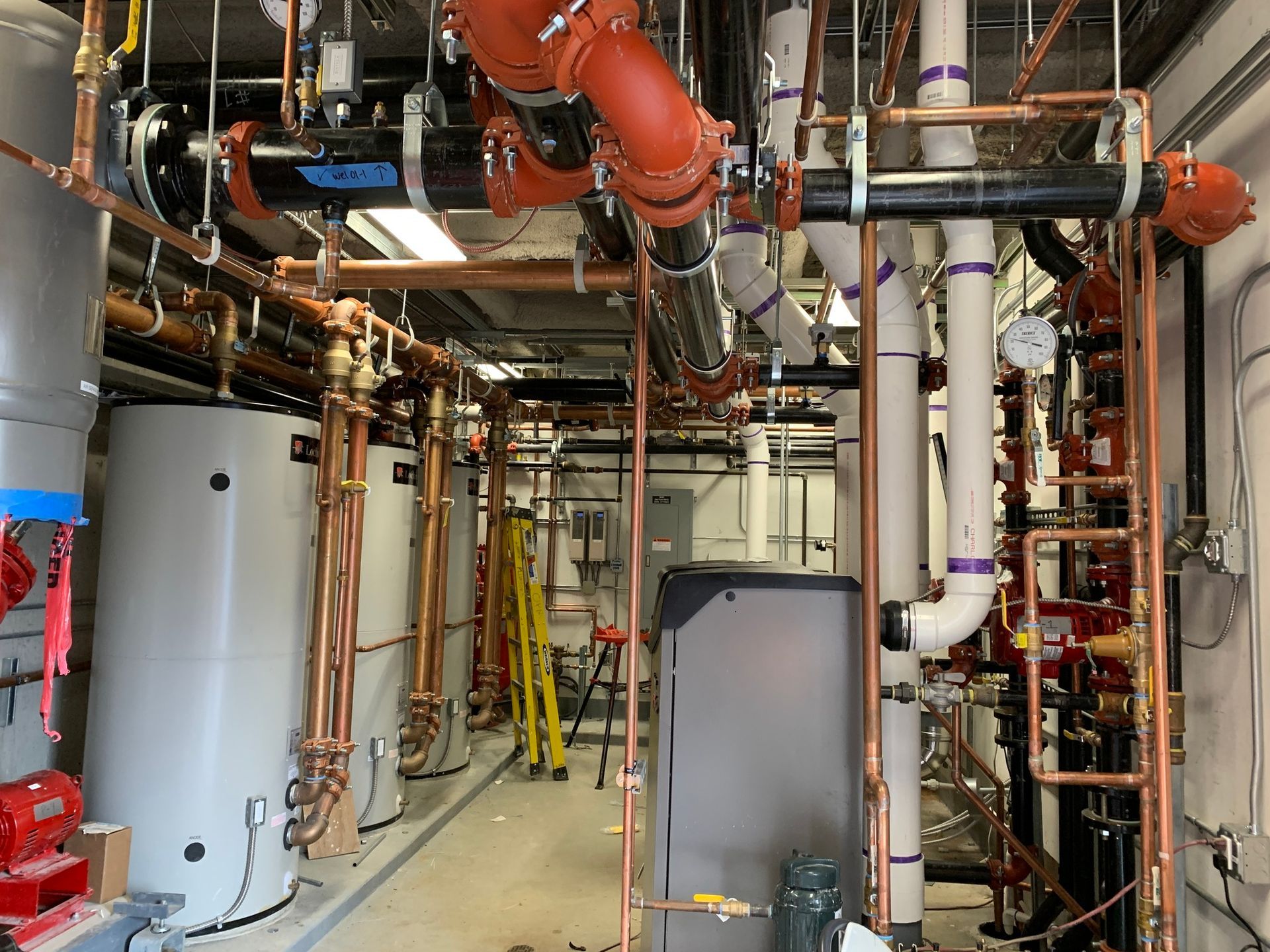 Mechanical room with multiple tall gray water heaters, complex copper piping, and industrial black and orange fittings.