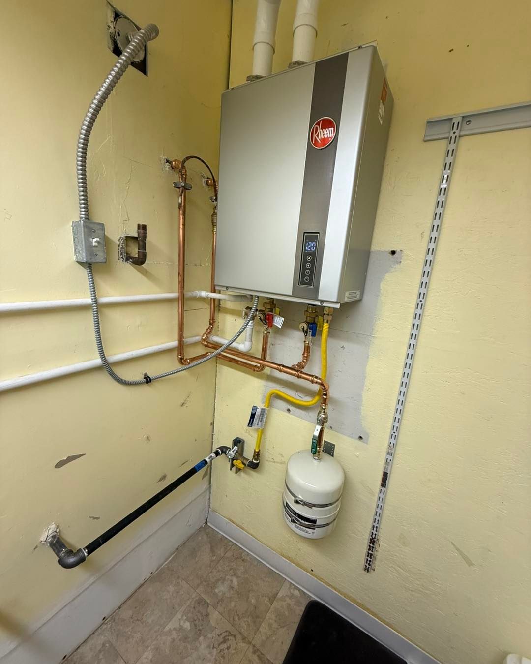 Rheem tankless water heater mounted on a yellow wall with exposed copper plumbing, a gas line, and a thermal expansion tank.