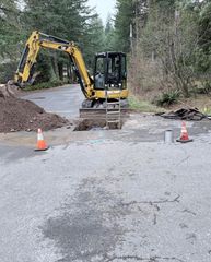 A yellow excavator sits by a hole in a paved area, marked by orange safety cones and a ladder near a forest edge.