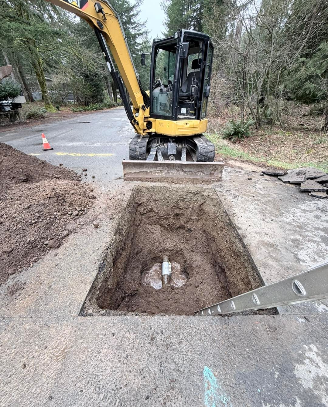 A yellow excavator parked behind a rectangular excavation in an asphalt driveway with a metal ladder inside the hole.