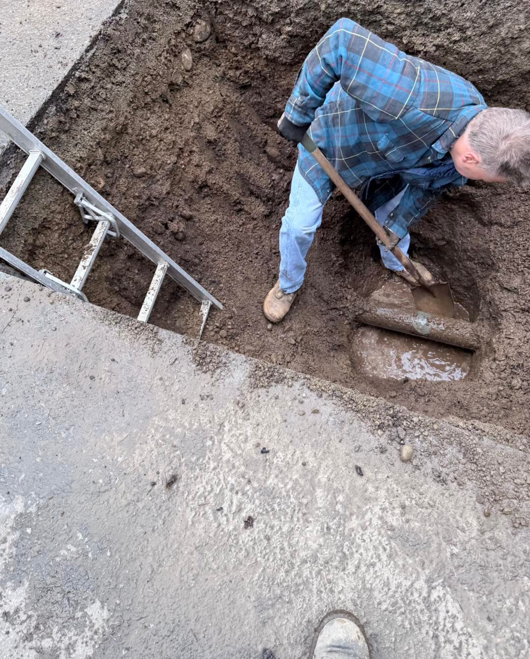A person in a blue plaid shirt uses a shovel to clear dirt around a pipe at the bottom of an excavation pit.