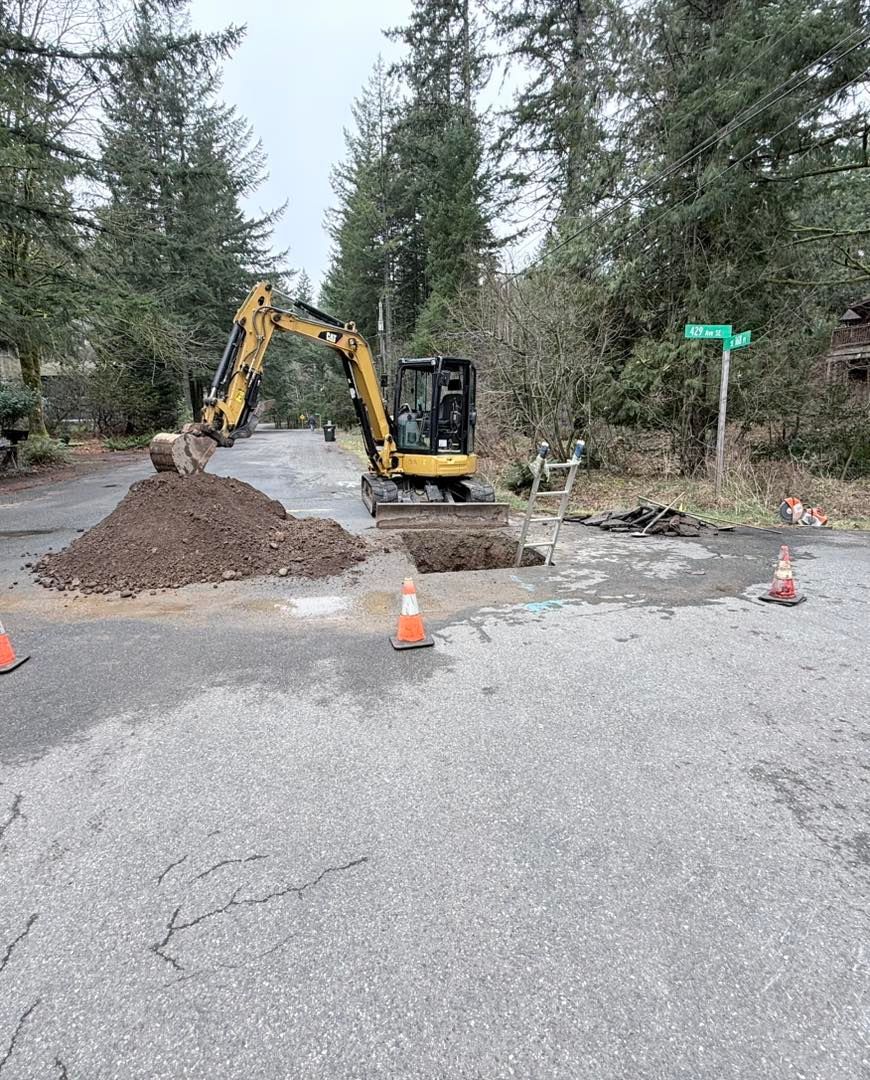 A yellow excavator works on a small road trench near a pile of dirt and traffic cones in a wooded area.