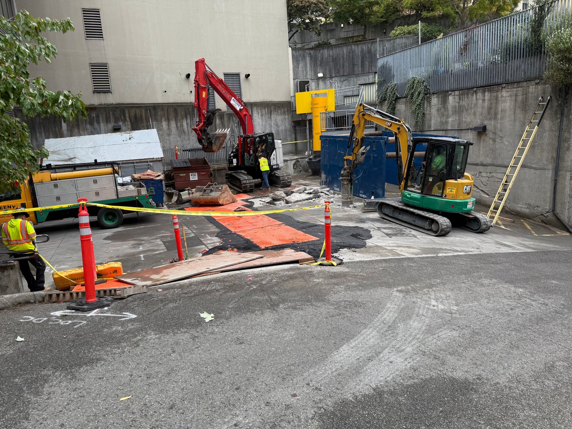 Construction site with a red excavator, a yellow compact excavator, a worker in safety gear, and orange safety cones.