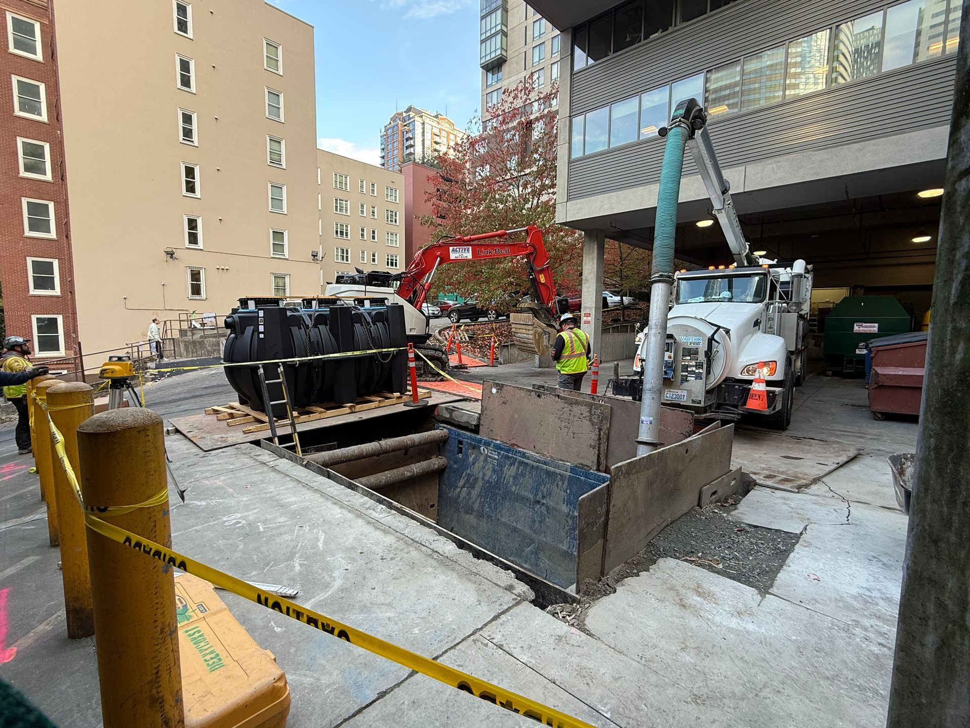A construction site with an excavator, a vacuum truck, and a deep trench being worked on by a person in a safety vest.
