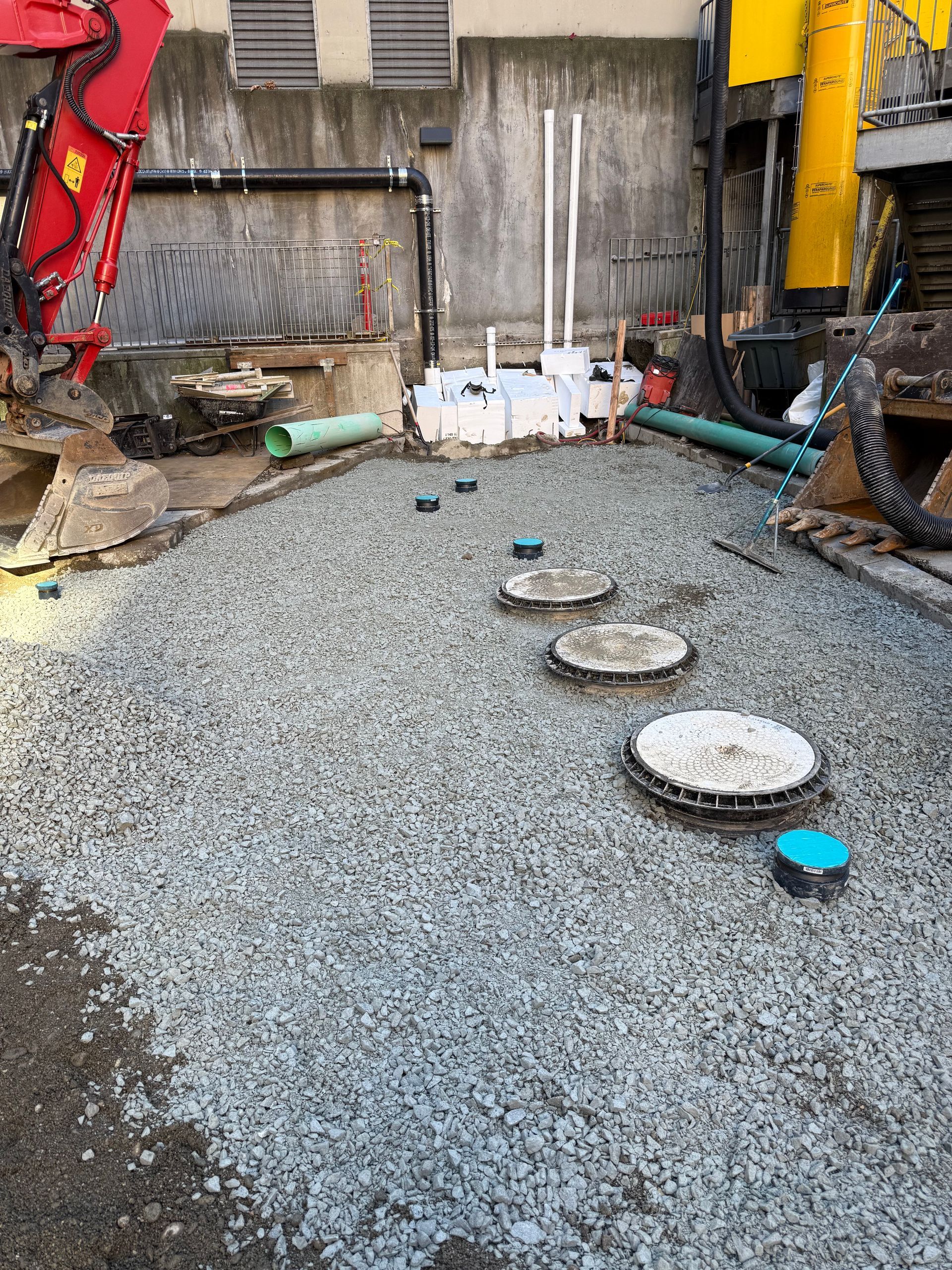 Construction site showing a gravel-covered area with several circular access manhole covers and pipes near machinery.