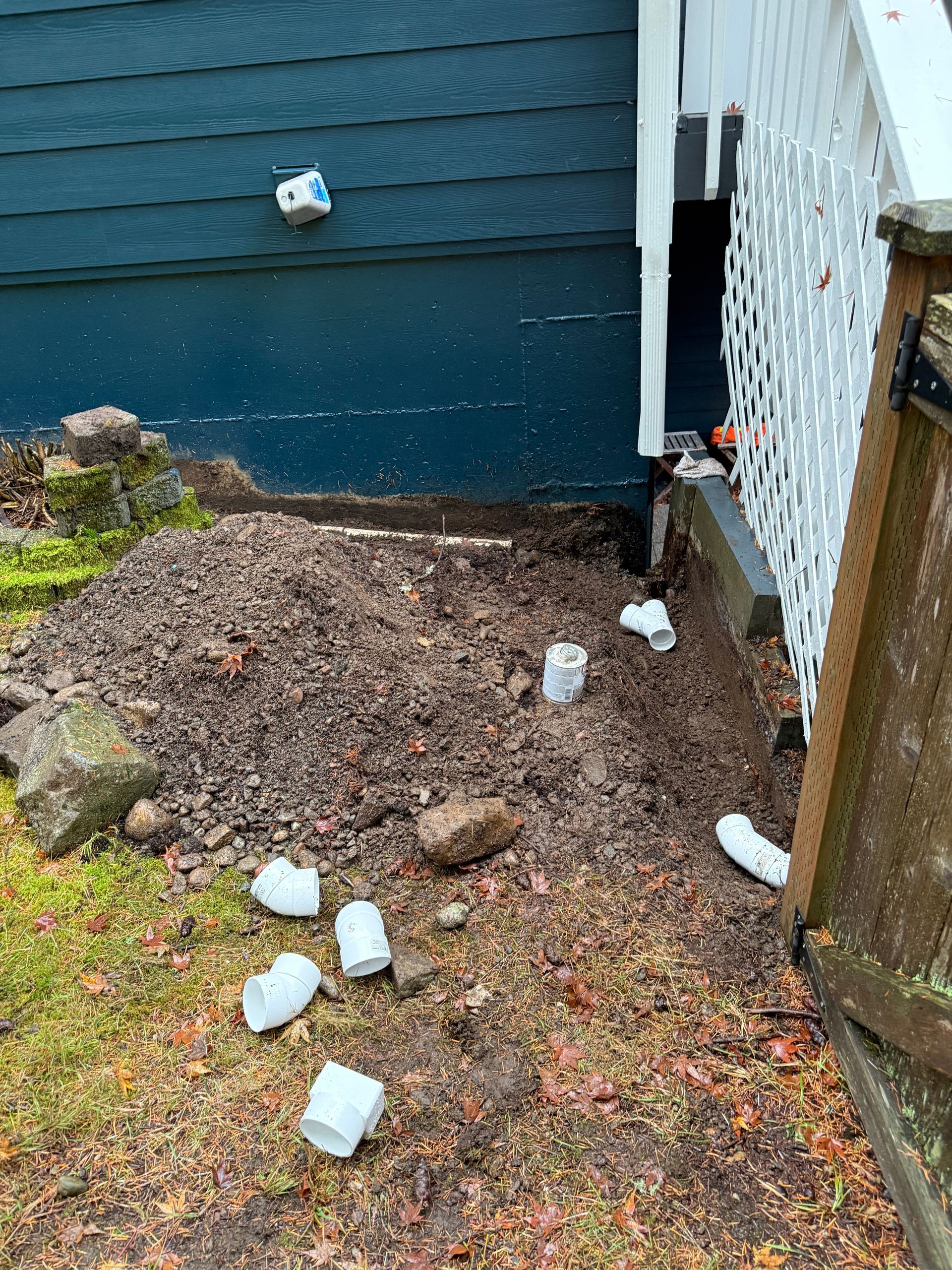 A pile of dirt and several scattered white PVC plumbing fittings next to a blue house and white lattice deck skirting.