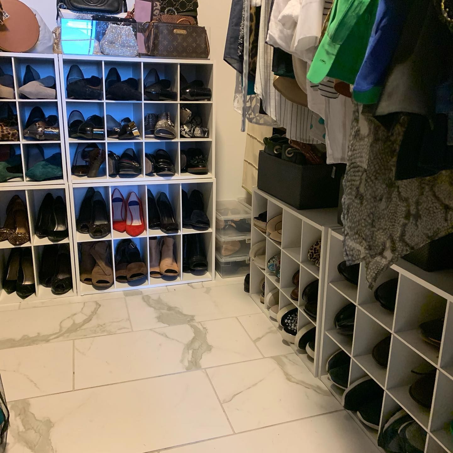 A walk-in closet with white shelving units filled with shoes and bags. Marble tile flooring.