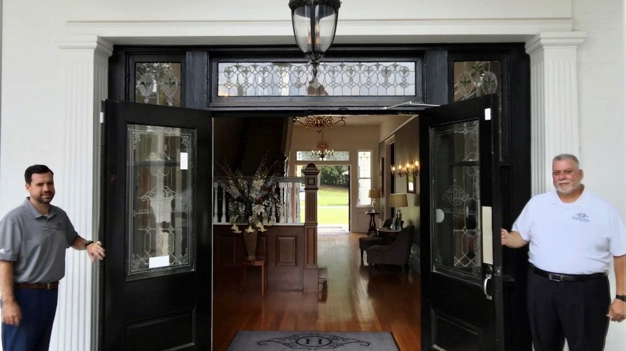 Two people stand at the open entrance of a building with black doors, leading into a lobby with wooden floors.