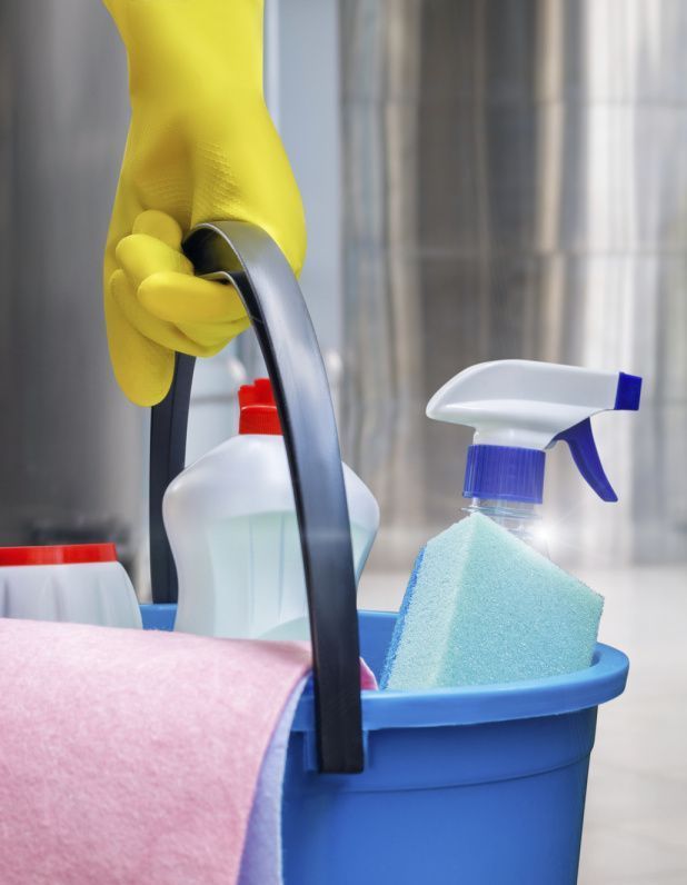 Hand in yellow glove holds a blue bucket with cleaning supplies: sprays, sponge, and rag.
