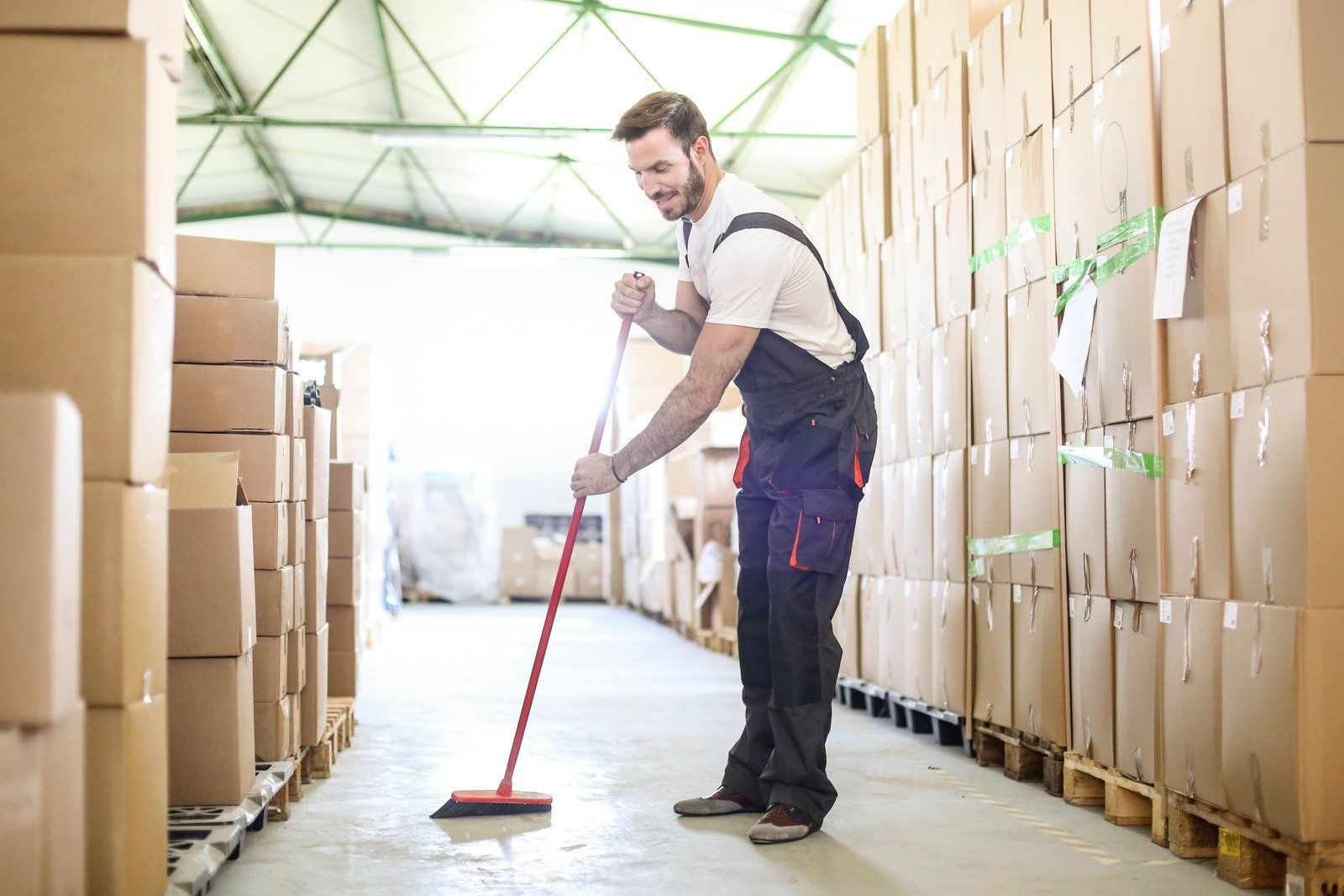 Man sweeping warehouse floor with red broom; boxes stacked on either side.