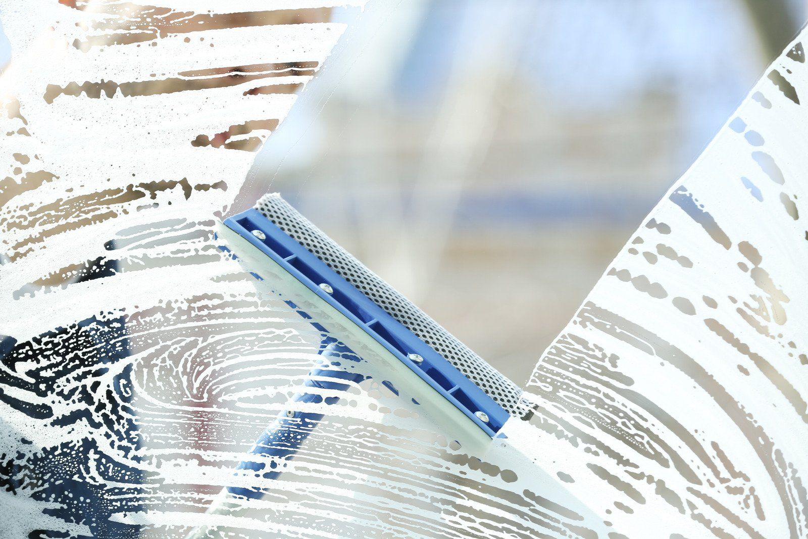 Squeegee cleaning a soapy window pane, leaving streaks. Close-up, daytime, blue and white tool.