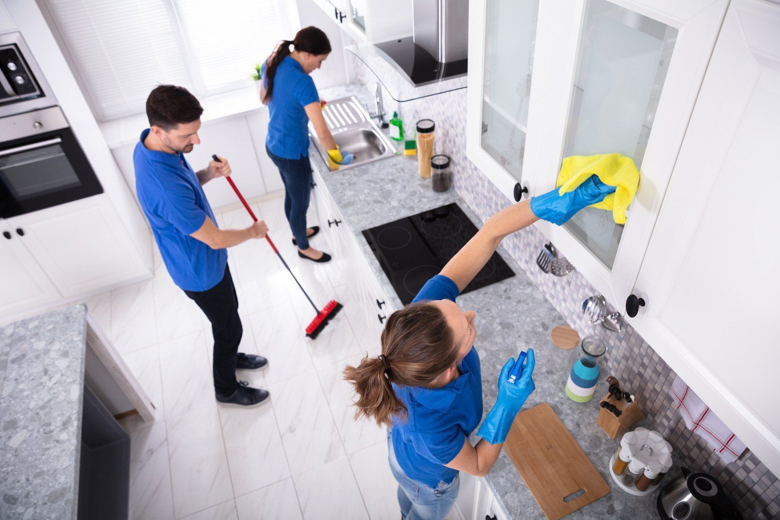 Three people cleaning a kitchen; one sweeping, one wiping the sink, and one wiping a cabinet.