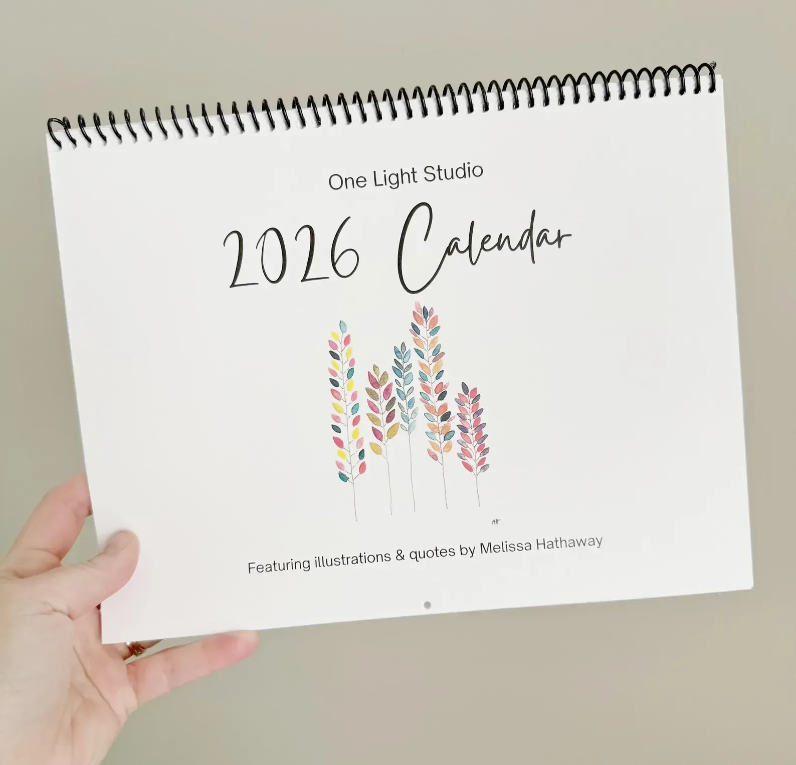 Hand holding a 2026 calendar with a spiral binding and colorful leaf design on a beige background.