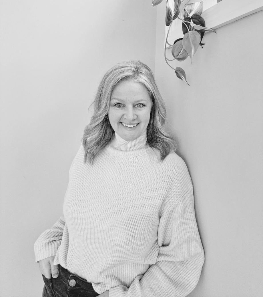 Woman in white sweater smiles, leaning against wall near a plant.