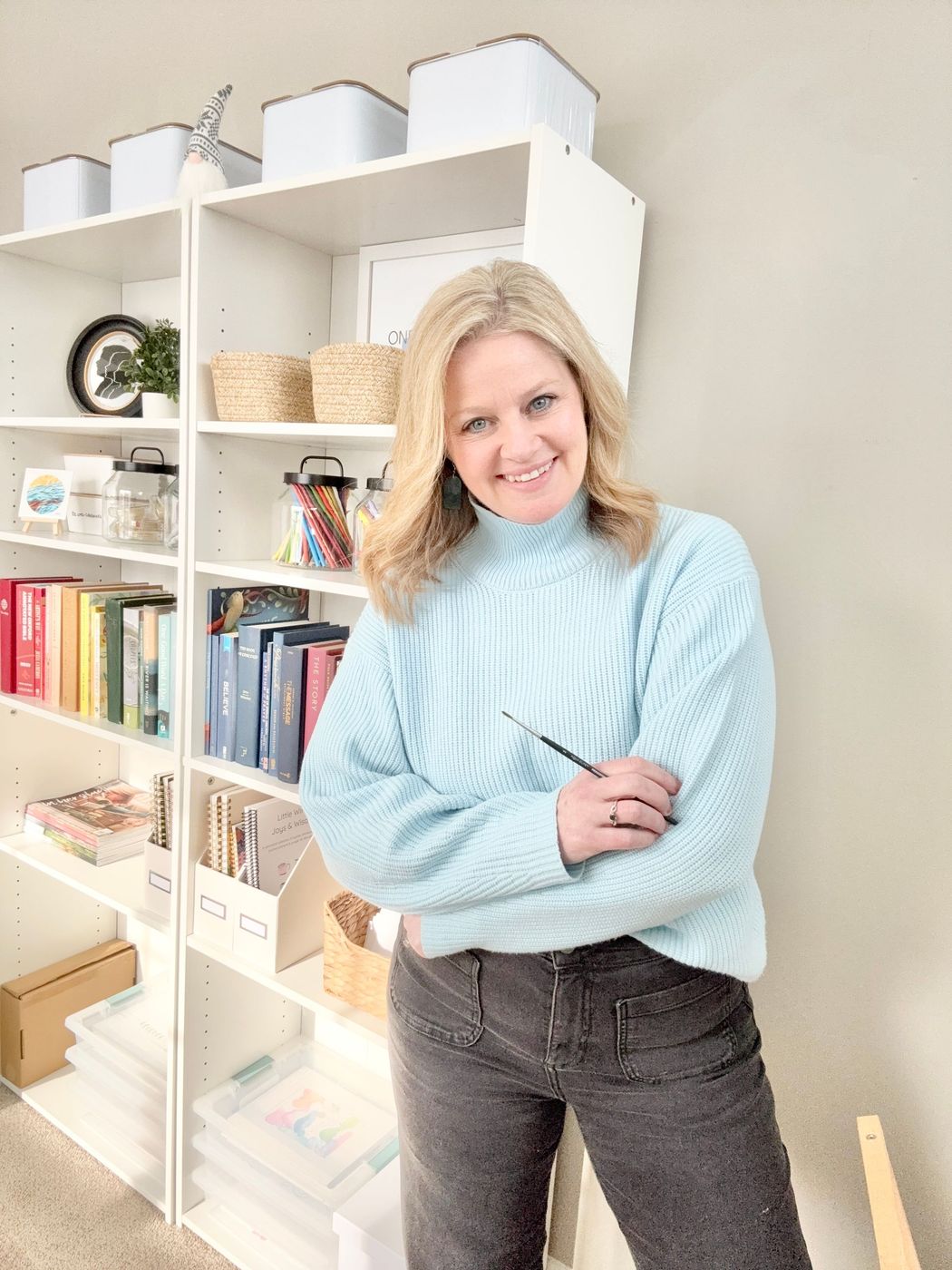 Woman smiling, wearing a blue sweater, standing in front of white bookshelves with storage containers and books.