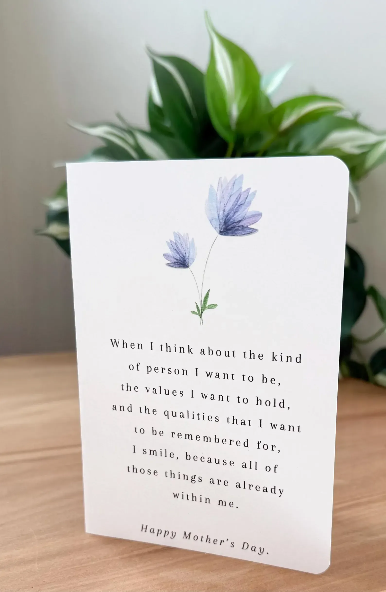 White greeting card with purple flowers and handwritten message in front of a green plant on a wooden table