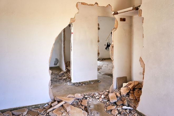 Vista interna di un edificio danneggiato, pareti con grandi buchi, detriti sul pavimento, vista su altre stanze.