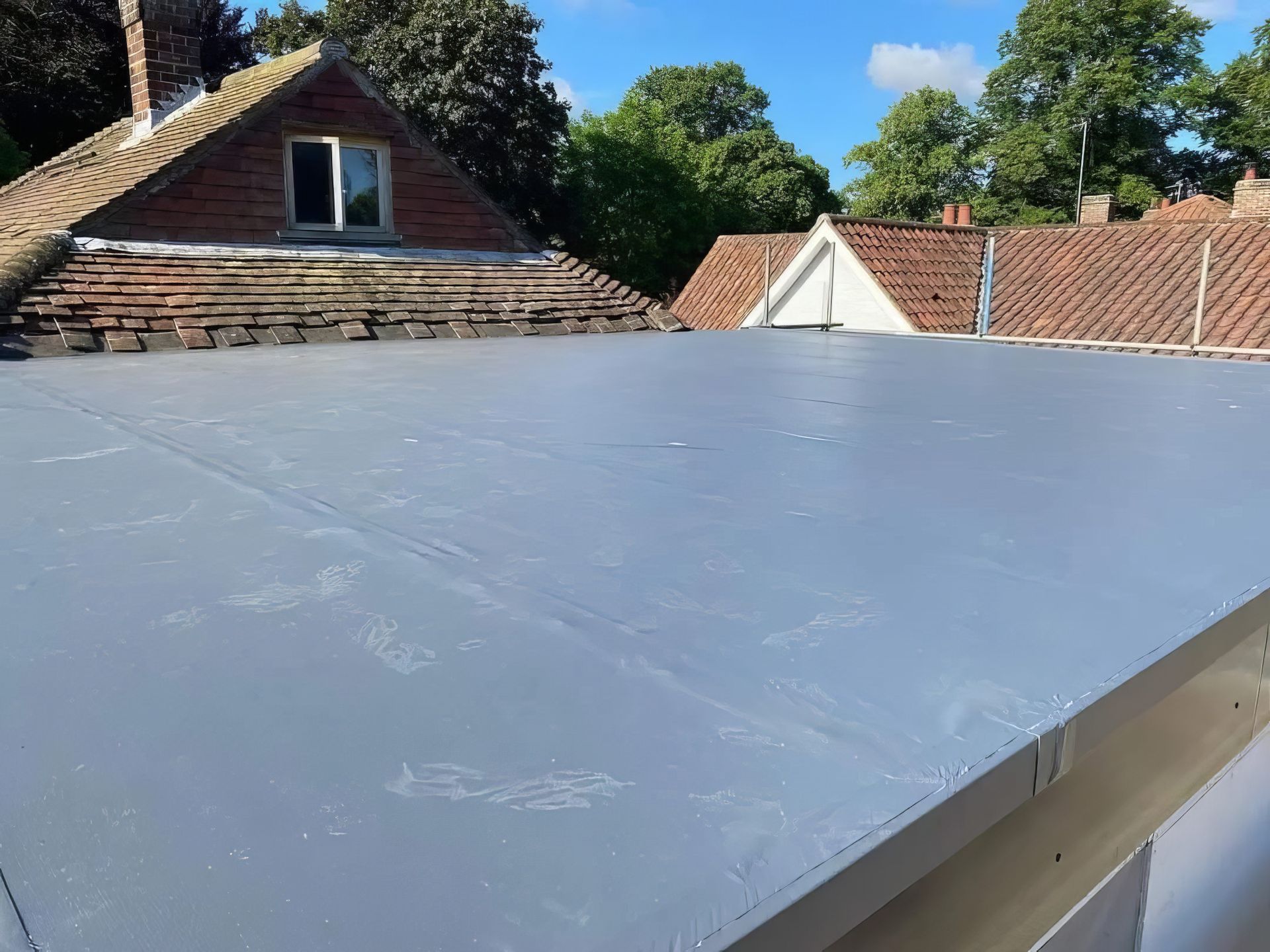 Flat gray rooftop membrane with brick houses and trees in the background under a blue sky