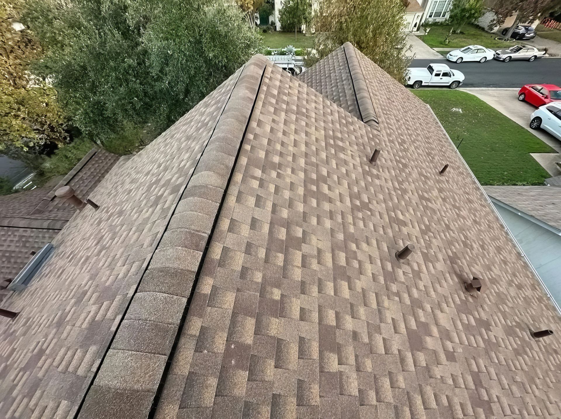 Aerial view of a shingled house roof with trees and parked cars below