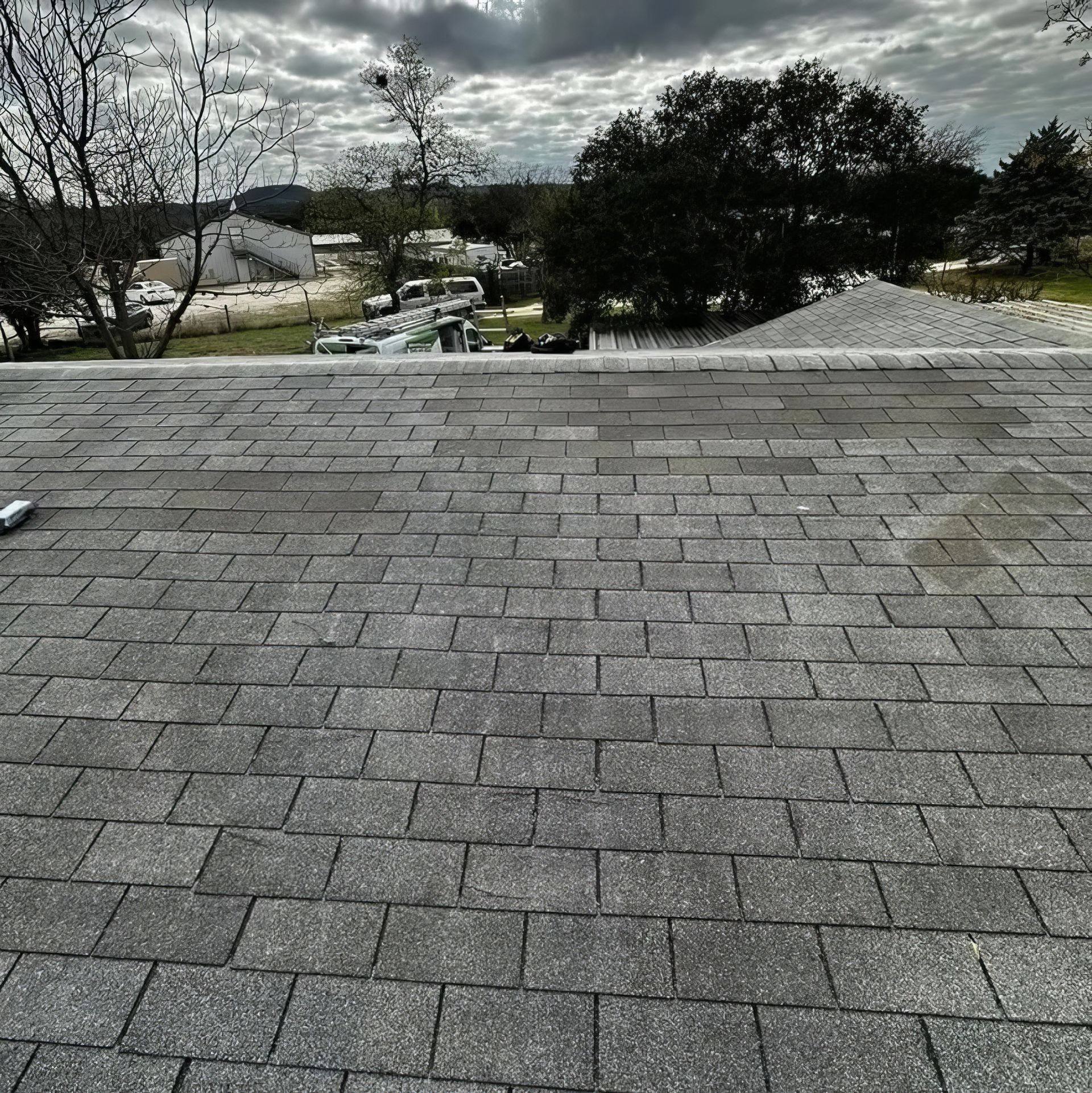 Rooftop shingles in foreground under a cloudy sky, overlooking trees and a residential area.