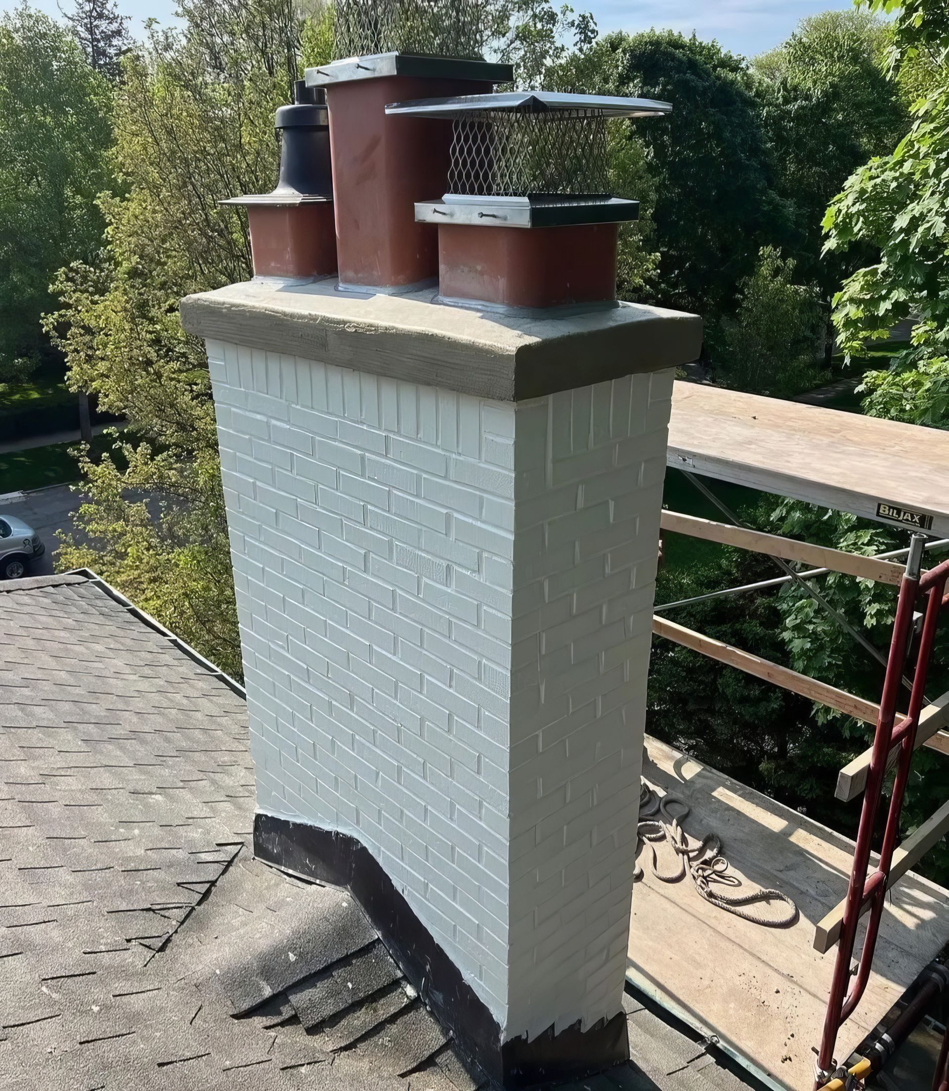 White brick chimney with red metal flues on a rooftop surrounded by trees