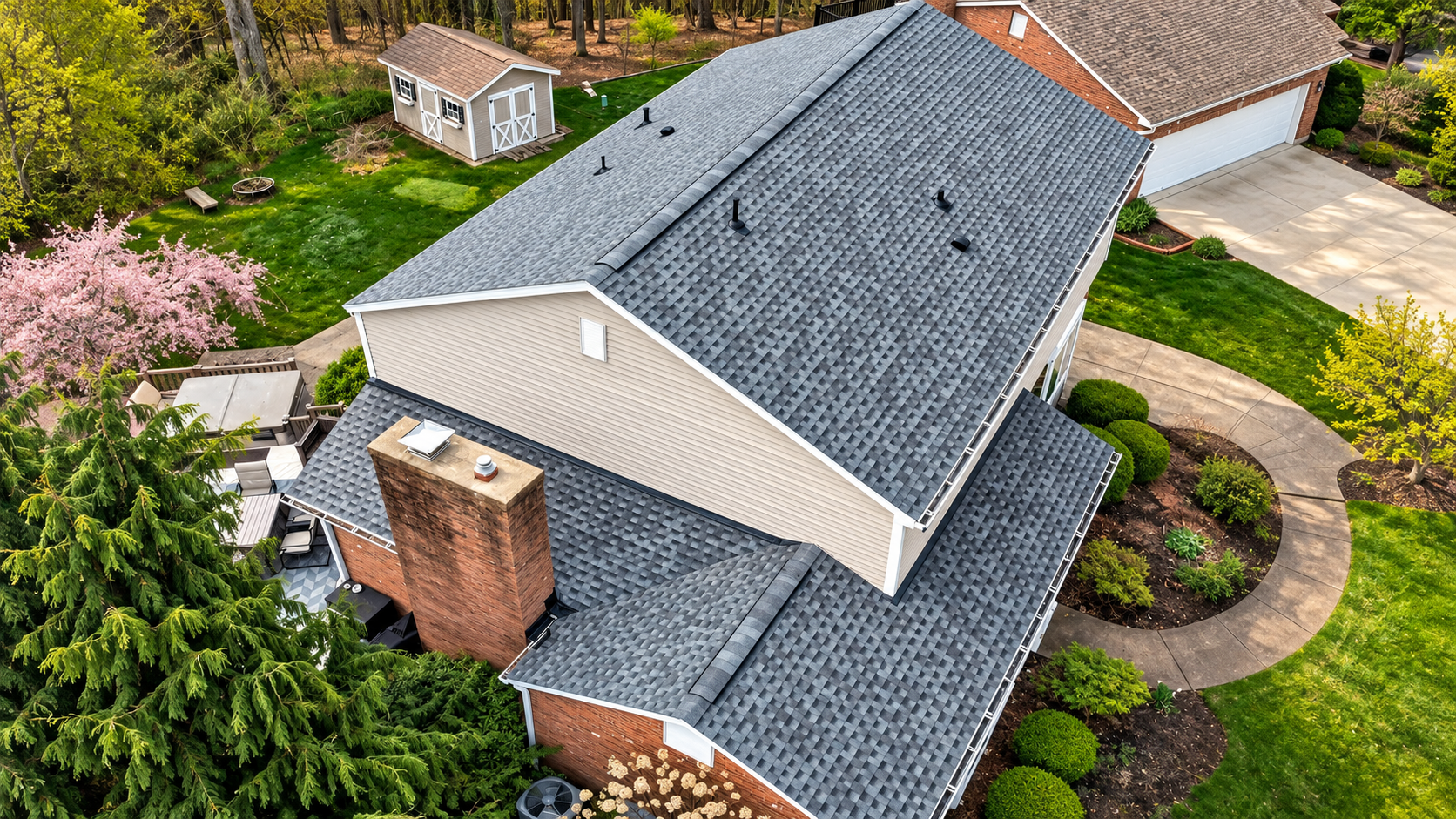 Aerial view of a house with a gray roof, brick chimney, and landscaped yard, plus a small shed nearby