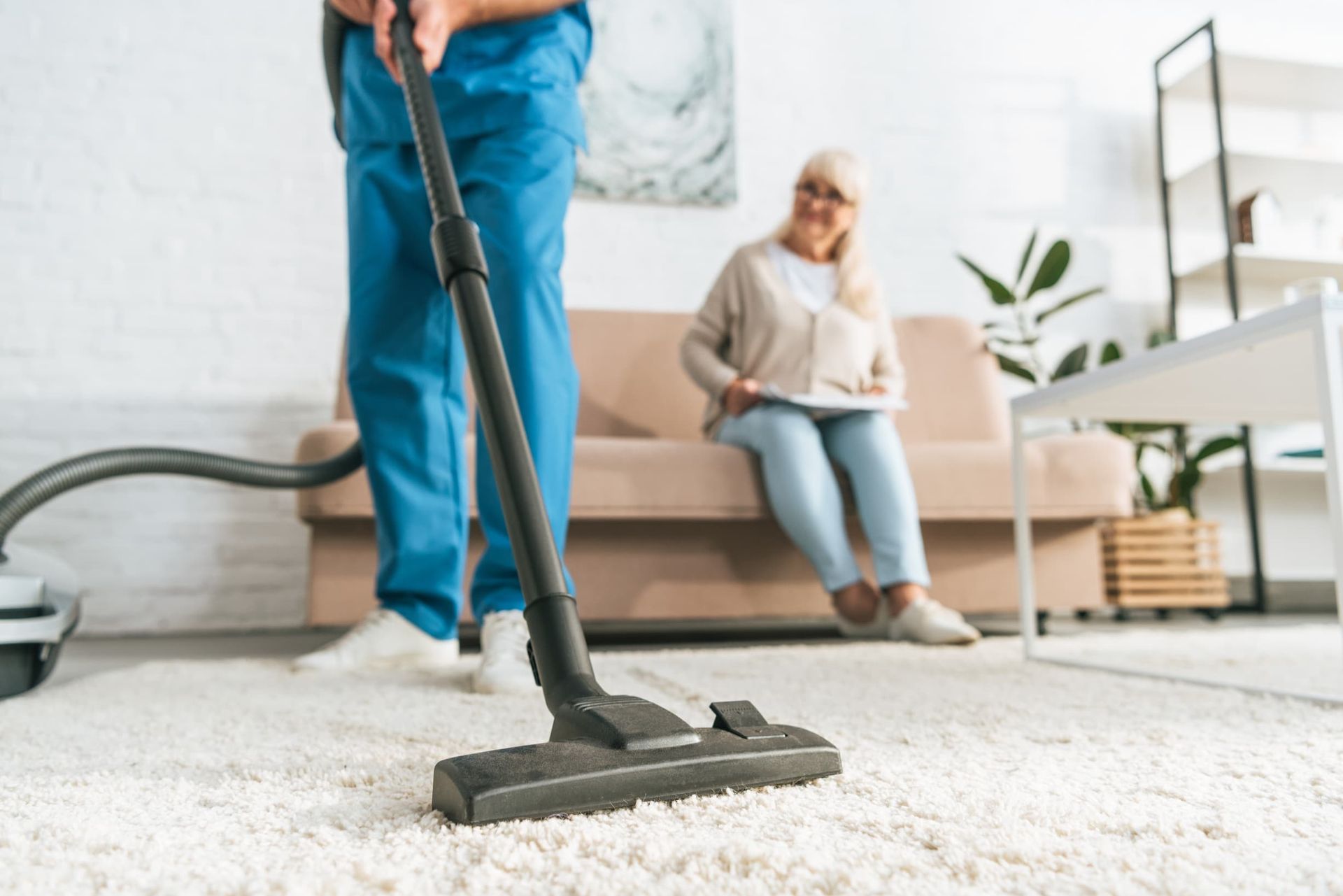 Vacuuming a White Carpet, with An Older Woman Sitting on A Couch — Elder Care at Home in Southport, QLD