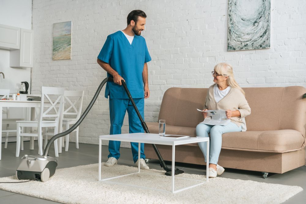 Man in Scrubs Vacuums in a Living Room While an Older Woman Sits on a Couch — Elder Care at Home in Highland Park, QLD