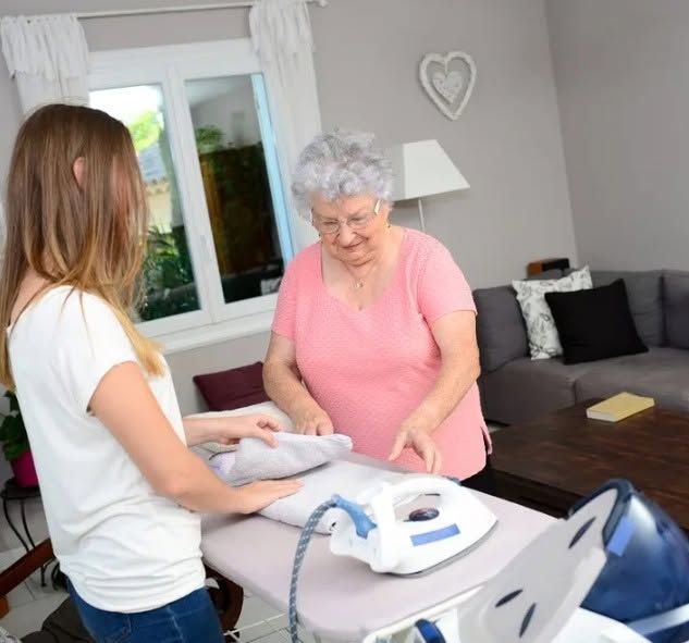 Man in Scrubs Vacuums in a Living Room While an Older Woman Sits on a Couch — Elder Care at Home by Andersen and Morgan in Highland Park, QLD