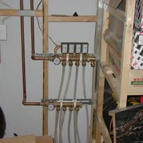 Copper pipes and manifolds with clear tubing, part of a radiant heating system, mounted on wood framing.