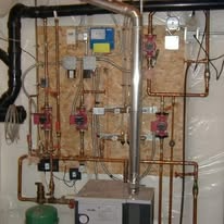 Complex copper plumbing system with pumps, valves, and a boiler, mounted on a wooden panel.