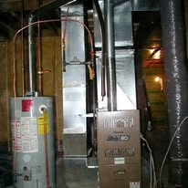 A furnace and water heater in a basement with silver ductwork, copper pipes, and exposed wires.