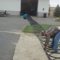 Two people installing black tubing on concrete, near a building with a large opening and a green lawn.