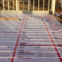 White pipes laid out across a foundation, likely for radiant floor heating, with a wooden frame structure in the background.