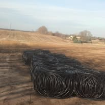 Coiled black irrigation tubing in a field, with a distant treeline and a partly cloudy sky.
