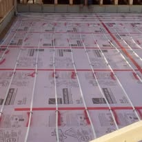 Piping laid in a grid pattern on pink insulation, likely for radiant floor heating, in a construction setting.