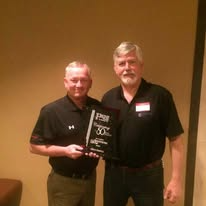 Two men holding a plaque, smiling. One in a black polo shirt, other in black shirt. Wooden background.