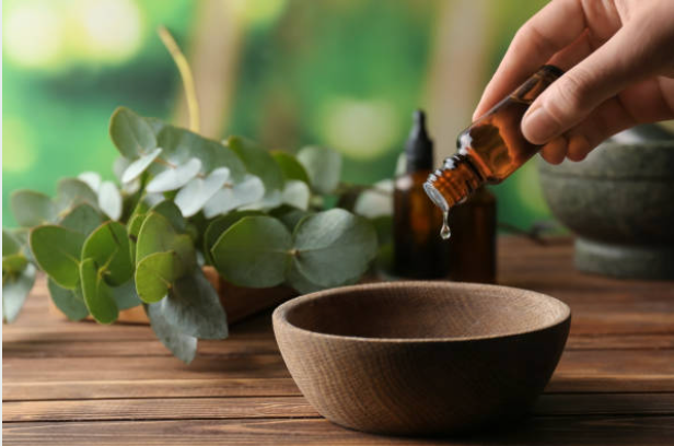 Met de hand druppelt men etherische olie uit een bruine fles in een houten kom, naast eucalyptusbladeren.