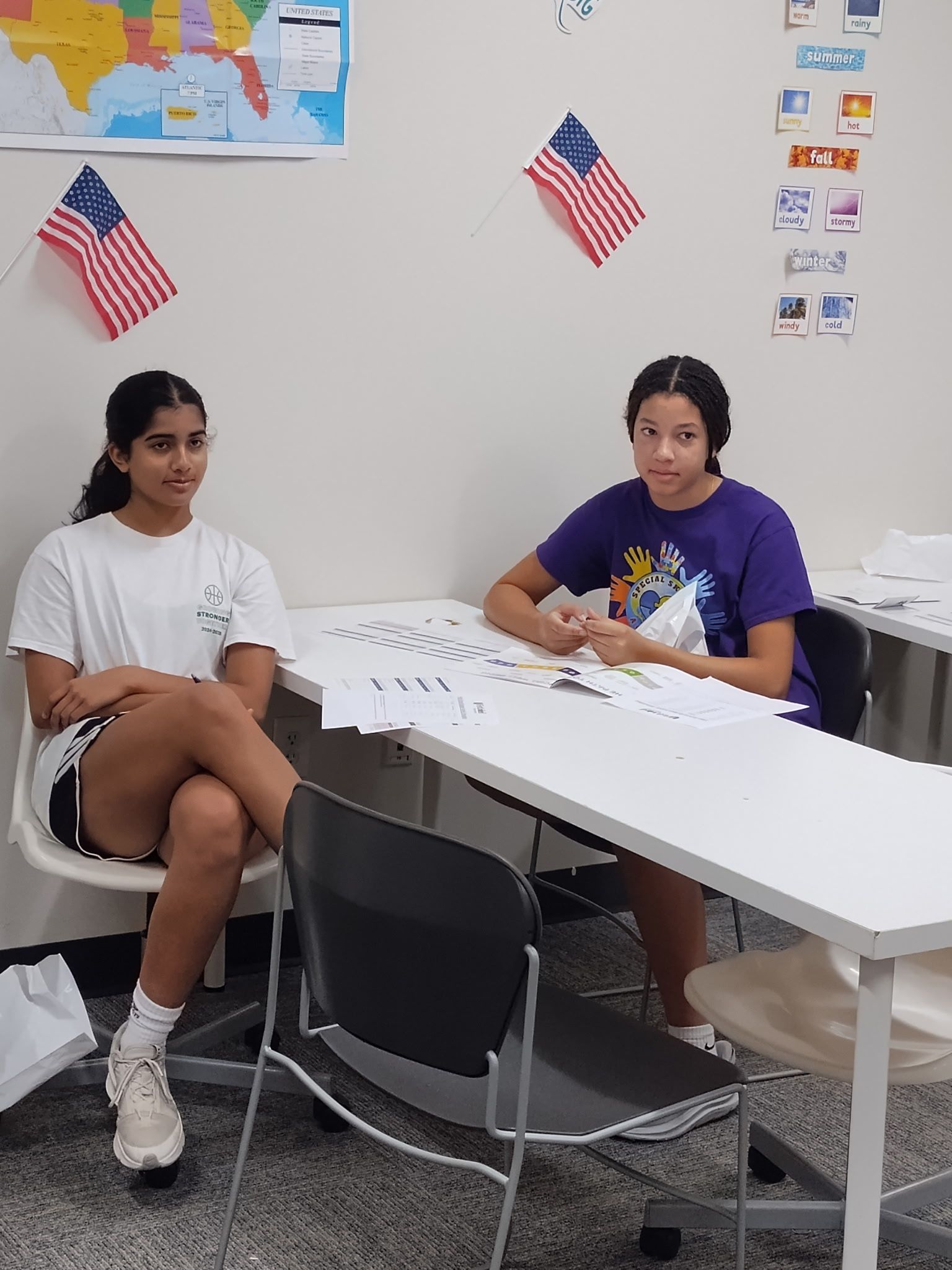 Two girls at a white table, possibly discussing papers. American flags are hanging on the wall.