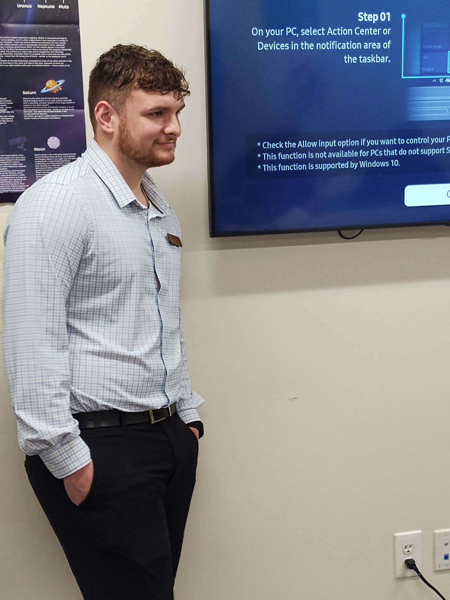 Man standing, smiling, hands in pockets, beside a screen displaying text. He's in an office setting.