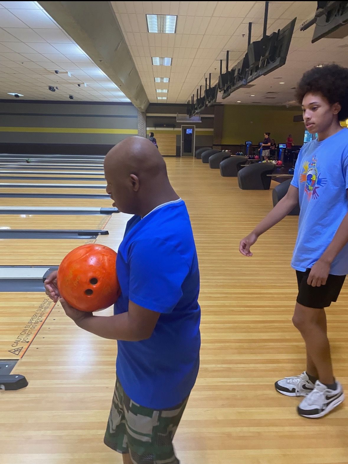 Man in blue holds a bowling ball, looking at the lane. A teen watches. Bowling alley.