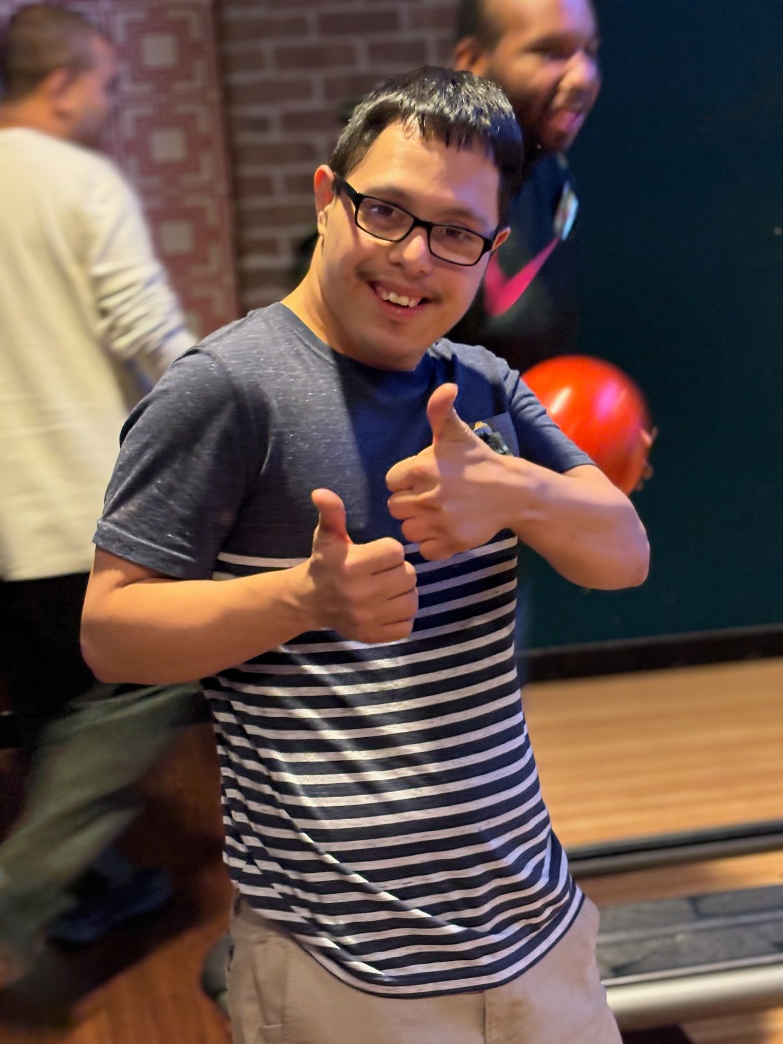 Man with glasses giving double thumbs-up at a bowling alley. He wears a striped shirt and smiles.